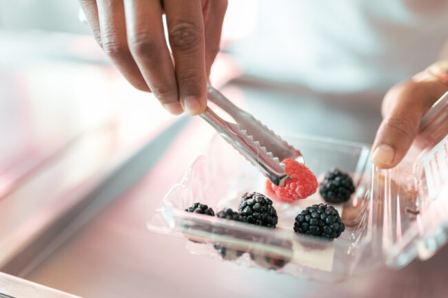 An ice-cream parlor worker is preparing some raspberry and blackberries on the roll machine using tongs
